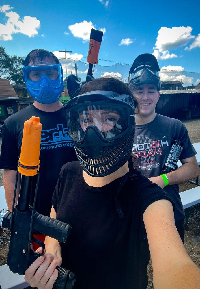 Three people wearing protective masks and holding paintball guns stand outdoors in front of picnic tables & a bright blue sky