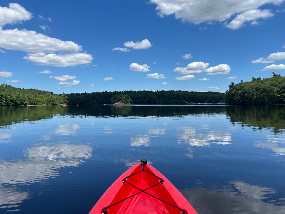Lake view from a red kayak: a dark green line of trees crosses the horizon in the distance while fluffy white clouds drift across the blue sky, reflecting in the water ahead.