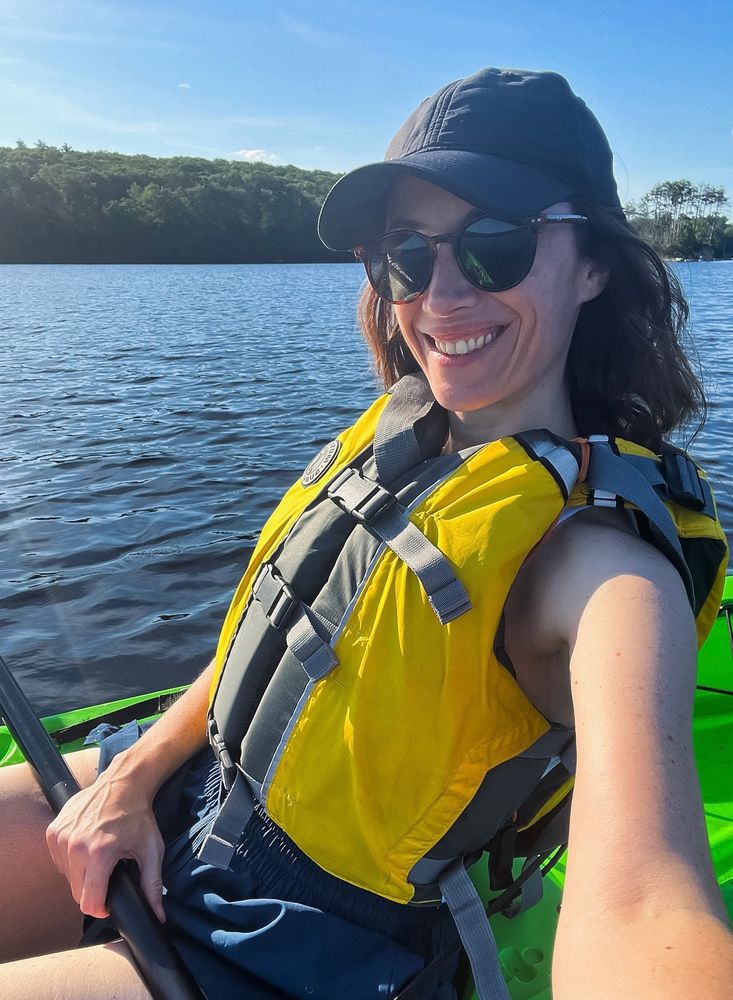A smiling woman wearing a bright yellow life jacket sits on a green kayak on the water 