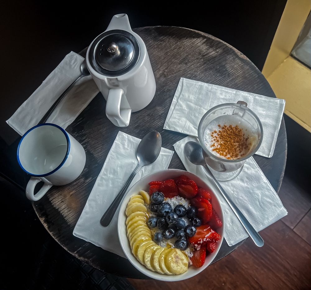 View of a small circular table top with napkins and spoons laid out, set with an açaí bowl topped with coconut & sliced fruit, a glass of cappuccino, a white & silver pot of tea and an empty white mug. The wood of the table blends in with the floor and walls around it, while the edge of a yellow window frame can be seen off to the right.