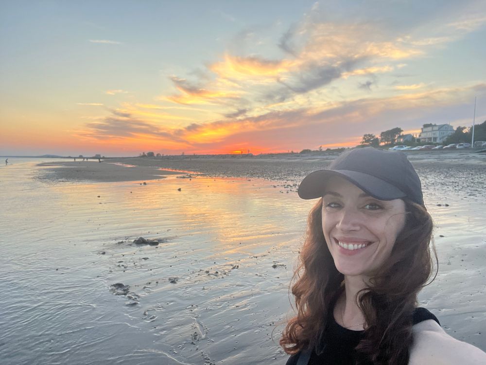 Photo of a smiling woman in front of a sunset beach view with pastel blue, yellow, & orange stretching across the horizon and reflecting on the wet sand