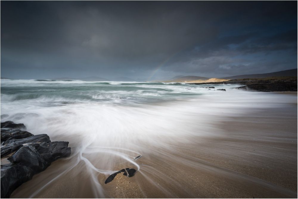 A small pebble interrupts the backwash on a Hebridean beach. beyond stormy seas lead the eye into a moody sky, a rainbow and the mountains beyond. 