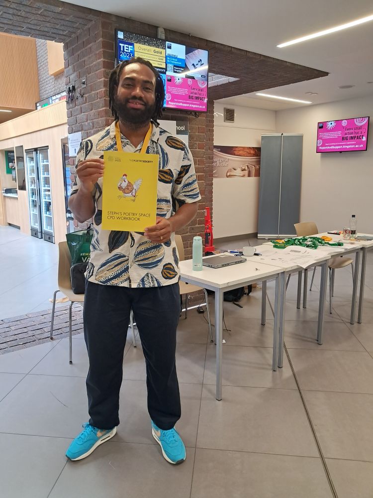 Photo of poet Ty'rone Haughton wearing a patterned shirt, black trousers and blue trainers. He is at the NATE conference and holds a yellow Steph's Poetry Space booklet.