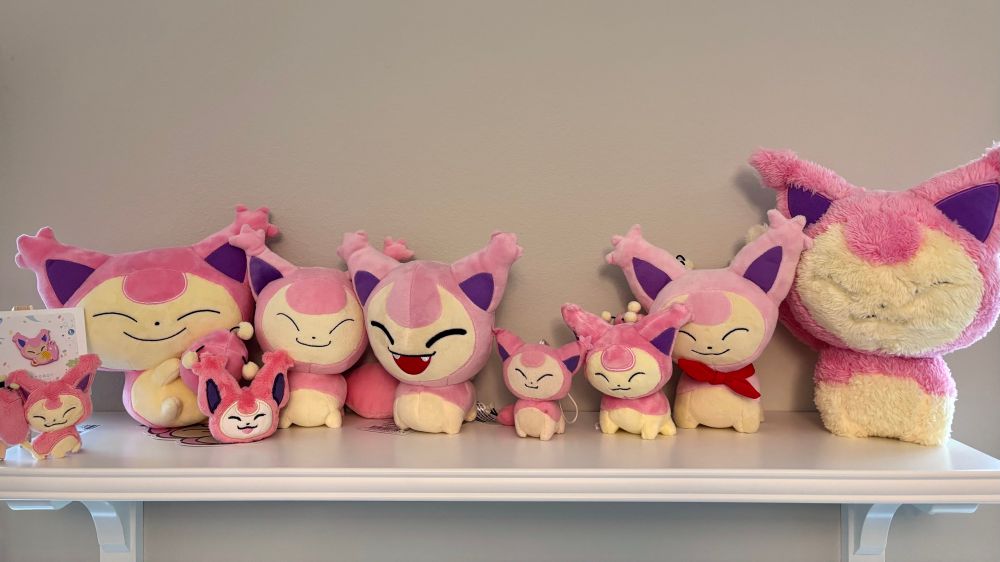 A photo of a mounted wall shelf full of plushes of the cat Pokémon Skitty. There are seven officially licensed Skitty plushes of varying sizes lined up across the shelf as well as two artist-made plush keychains. On the far left of the shelf there is a tiny easel with a Skitty pin from artist Mamobot.