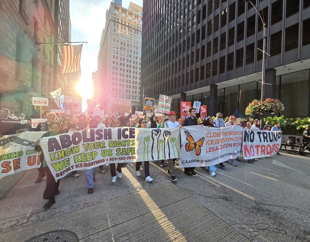 A protest for immigrant rights marching down a Chicago street past a black federal building- an American flag hangs from a skyscraper backlight by a glorious sunset