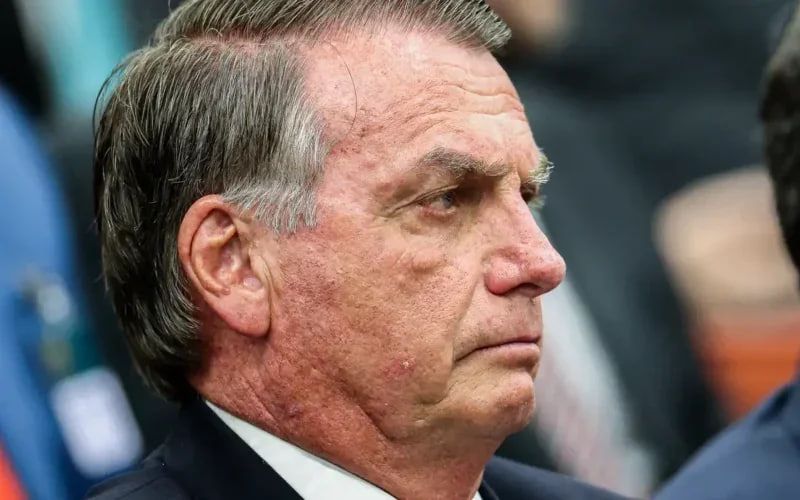 Close-up of Jair Bolsonaro, Brazil’s former president, seen in profile. He wears a dark suit jacket and white shirt. His expression is stern, with visible skin blemishes and marks on his face, under neutral indoor lighting.