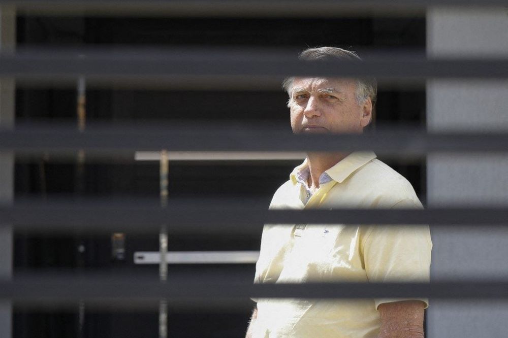 Former Brazilian president Jair Bolsonaro is seen standing behind a gate, wearing a yellow polo shirt and looking straight ahead. The horizontal bars of the gate give the impression he is behind bars. The photo was taken by Sergio Lima/AFP in front of the house where he is under house arrest.
