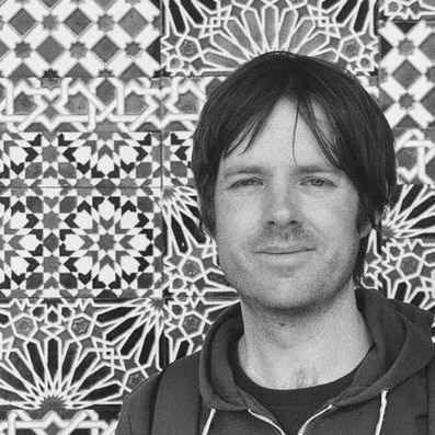 A man with dark hair parted in the centre smiles at the camera, with abstract art on ceramic tiles behind him. Black and white. Board game designer Matthew Dunstan.