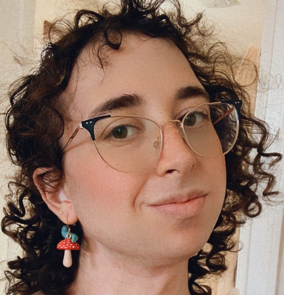 A woman with brown curly hair and glasses, wearing mushroom earrings, smiles at the camera. Marceline Leiman.