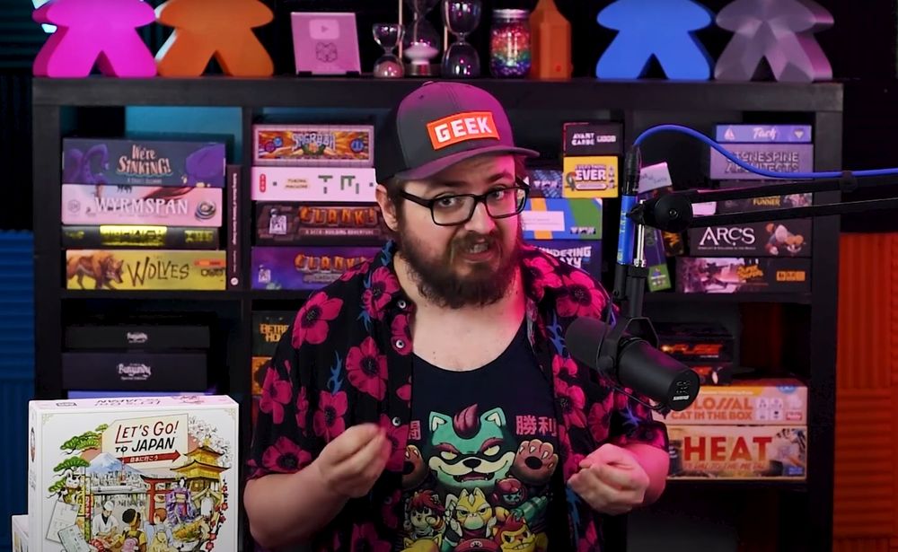 A man wearing a baseball cap which says geek speaks to the camera while sitting in front of a shelf of board games. He has a brown beard and is wearing a black open shirt with pink flowers on, and a black t-shirt featuring a teal-coloured bear-like character. A copy of the board game Let's Go! To Japan is on the table to the left.