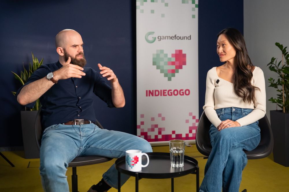 Gamefound CEO Marcin Swierkot, left, and Indiegogo CEO Julie dePontbriand. Swierkot, wearing a dark blue shirt and light blue jeans, gestures with both hands to emphasise a point he is making to dePontbriand. He has a shaved head and sports a thick brown beard. DePontbriand also sits, wearing a white long-sleeved top and blue jeans, and has long dark hair. Behind the pair is a vertical banner, white, which has a green and pink/red heart in its centre. Above is the Gamefound logo and below the words INDIEGOGO in pink.