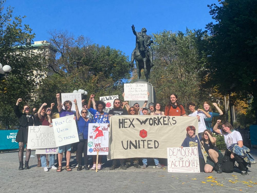 A group of about 20 people pose in front of a statue of a man on a horse, holding various signs promoting the union for workers of the Hex & Company board game cafe in New York.