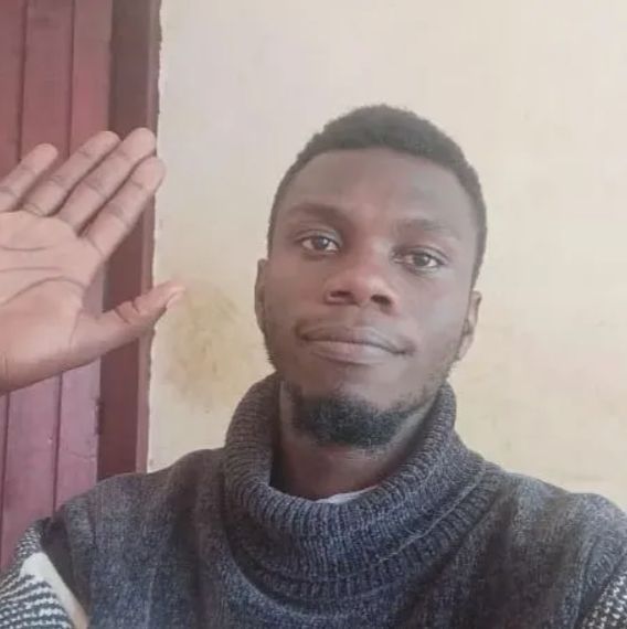 A close up of a man with short black hari and a chin beard, wearing a grey jumper, waving. Ashraf Braden.