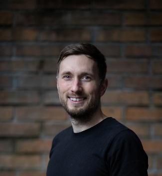 A man turns to smile at the camera with his teeth showing. Short dark hair swept to one side and a short beard. Wearing a black t shirt, in front of blurred red bricks in the background. Board game designer Tomas Holek.