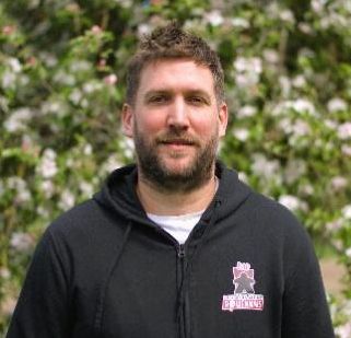 A bearded man with short, spiky brown hair looks directly into the camera. He wears a black hoodie with a meeple logo on one breast, and a white t-shirt underneath. Blurred foliage and pink flowers are visible behind. Board game designer Gregory Grard.