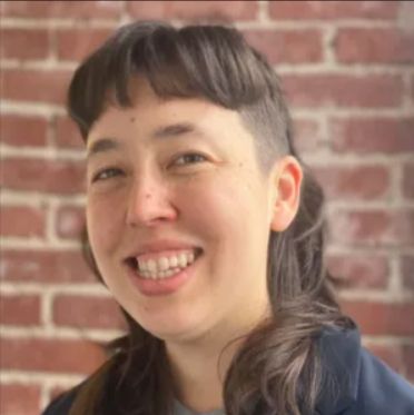 A woman with long brown hair, shaved at the side above the ears, smiles at the camera. Lyla McBeath Fujiwara.