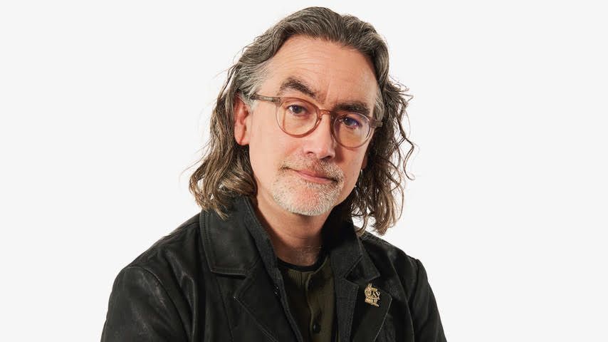 A man with long dark hair and glasses and a white goatee beard looks at the camera