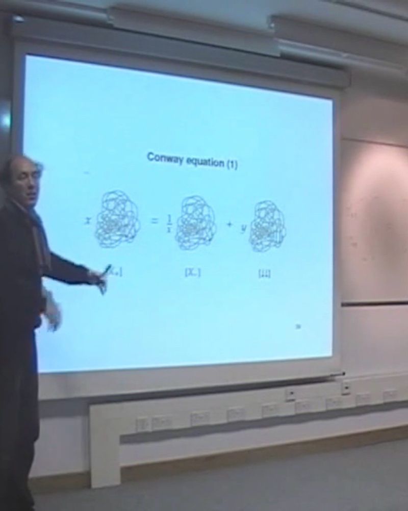 A wiry-haired french professor stands in front of a power point presentation pointing at three dense bundles of knots
