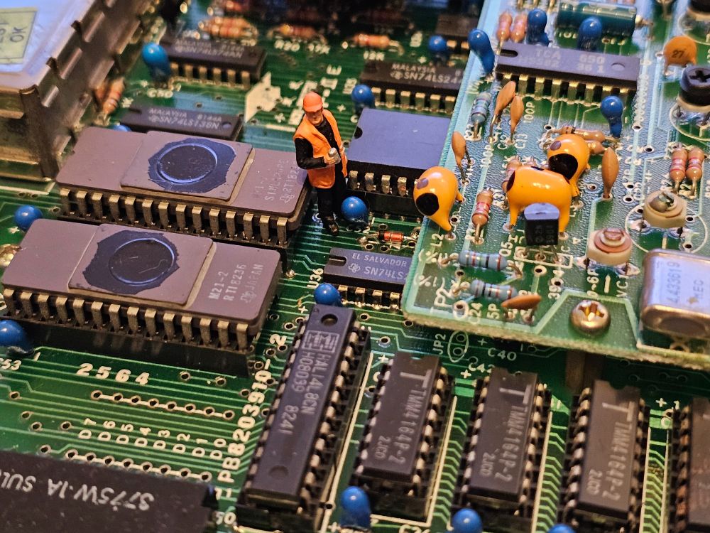 Close-up image of an 'unidentified' circuit board.  Standing amongst the computer chips, with a cup of tea, is a 1:76 scale figure in a fluorescent orange jacket and hard hat.