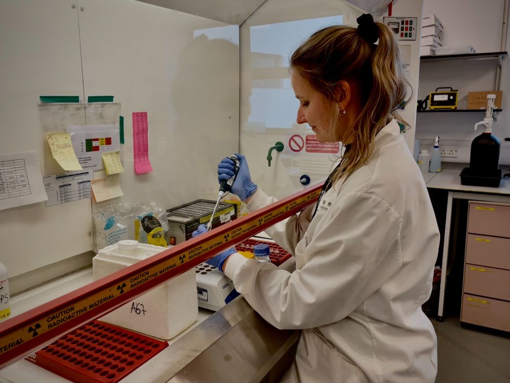 A woman in a lab space works with a pipette. Her arms are under a protective bar and she is surrounded by standard lab equipment.