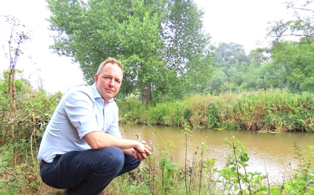 A photo of Richard Foord crouching on a riverbank looking toward the camera, with the river to the right of him. The river is brown in colour and is surrounded by grasses and trees