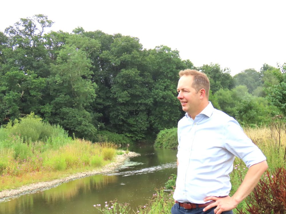 A photo of Richard Foord standing on a riverbank looking out onto the river to the left of him. He is standing with his hands on his hips, and the river itself is surrounded by grasses and trees. The water has a mirror sheen to it