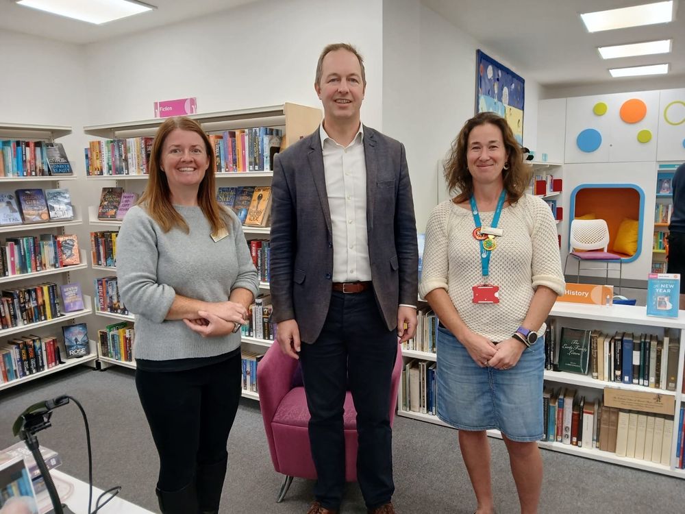 A photo of three people standing in a library, with Richard Foord in the middle and all three smiling toward the camera 