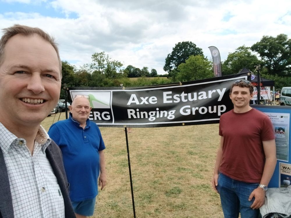 A selfie taken by Richard Foord on the left, with two men behind him either side of a black and white banner reading “Axe Estuary Ringing Group”. They are in a show ground 