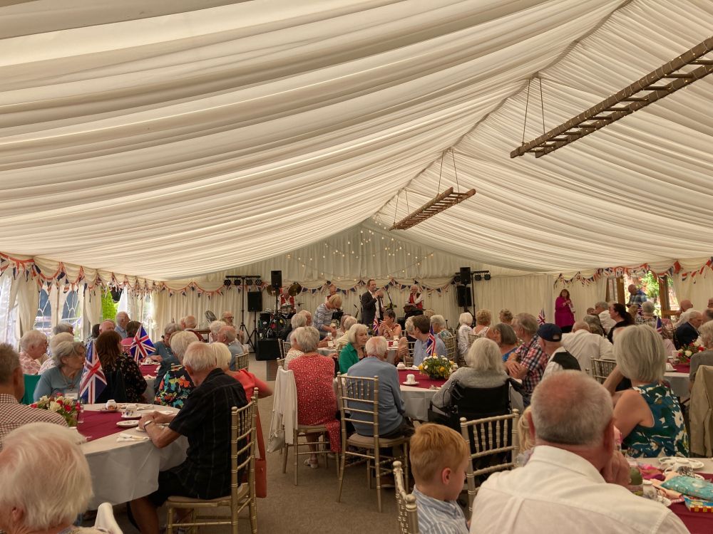 A photo of a group of people sat around tables in a large white tend. Around the tent is union flag bunting, and there are some lights strung at the far end. Richard Foord is standing at the far end with members of a jazz band where he is speaking into a microphone.