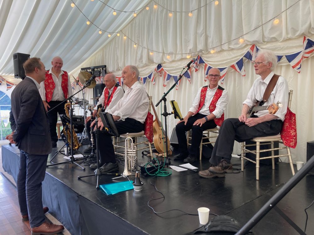 A photo of Richard Foord standing speaking to members of a jazz band on a stage in a large tent. The tent is white and there is union flag bunting on the back wall and lights strung from the ceiling. The jazz band has five members, four of whom are sitting on chairs, and the fifth is standing to the left of the other players. All are wearing white shirts, with three players wearing red waistcoats. There are also various instruments including guitars, a trumpet, a clarinet, and a tenor saxophone visible