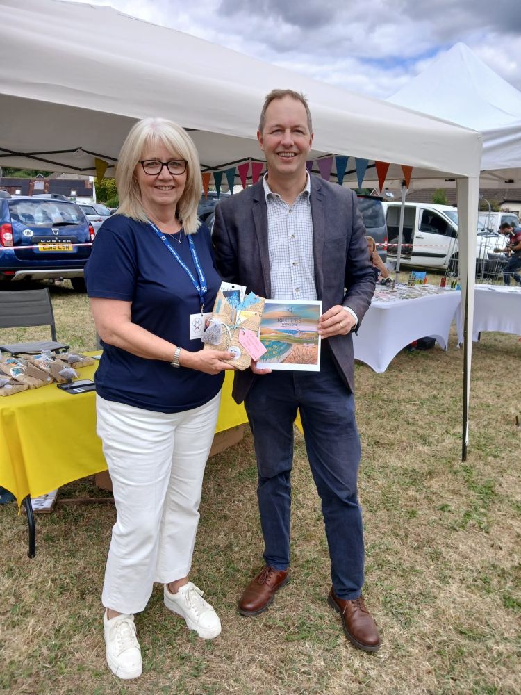 Richard Foord (right) standing at a stall in a showground with a woman to his left. Richard is holding a book with the title “Recipes with love”, and the woman is holding a package wrapped in brown paper