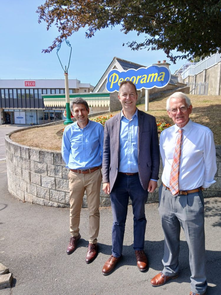 A picture of three men standing in front of a sign for Pecorama and a building with the PECO logo on it. In the middle of the three is Richard Foord. All three are looking toward the camera. The picture has been taken outside on a sunny day.