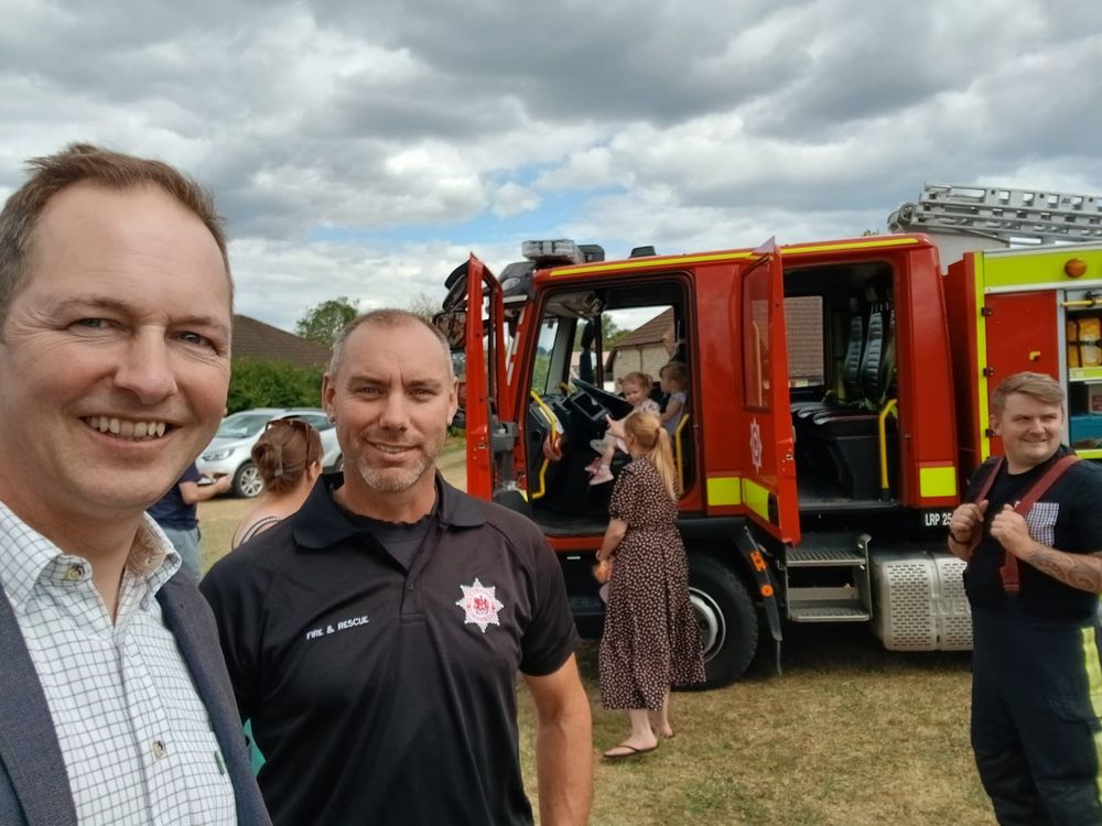 A selfie taken by Richard Foord on the left with a man in a fire brigade polo shirt. They are standing in a showground in front of a fire engine with its doors open 