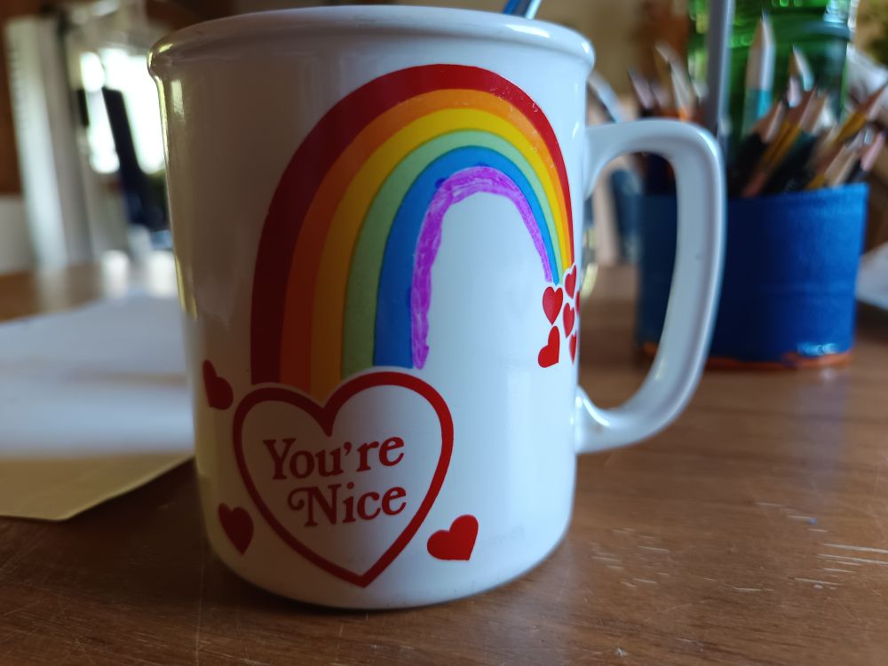 A white drinking cup with some red hearts on it. One big heart contains the words "You're Nice". Over it, there's a rainbow with only 5 colors (red, orange, yellow, green, blue) and a sixth stripe was sloppily added with purple sharpie