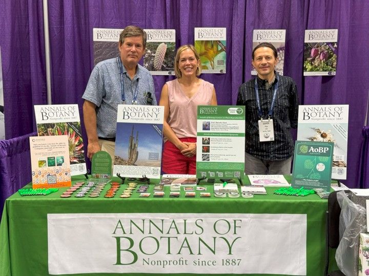 Rowan Sage, Rachel Shekar, and Xavier Picó standing behind the Annals of Botany booth at Botany 2025 conference. The booth displays journals, promotional materials, and signs for Annals of Botany, AoBP, and in silico Plants, all set against a purple curtain backdrop.