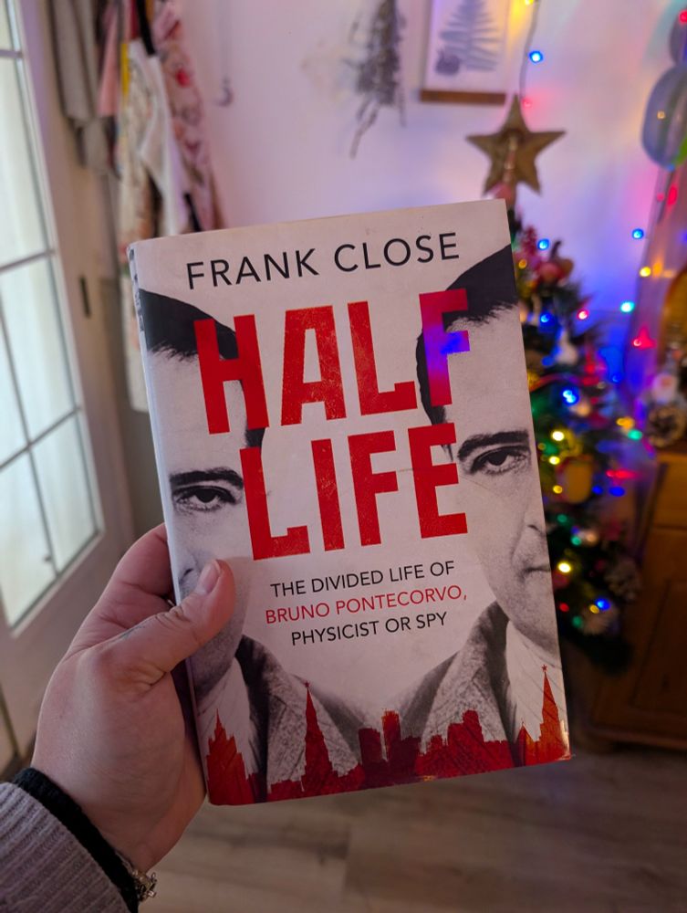 Sostengo en mi mano un libro grueso con sobrecubierta con grandes letras de estilo soviético "Frank Close HALF LIFE" en subtítulo con letra sin serifa " The divided life of Bruno Pontecorvo, physicist or spy". Al fondo hay un árbol de navidad pequeño con luces encendidas y mobiliario de cocina de bruja