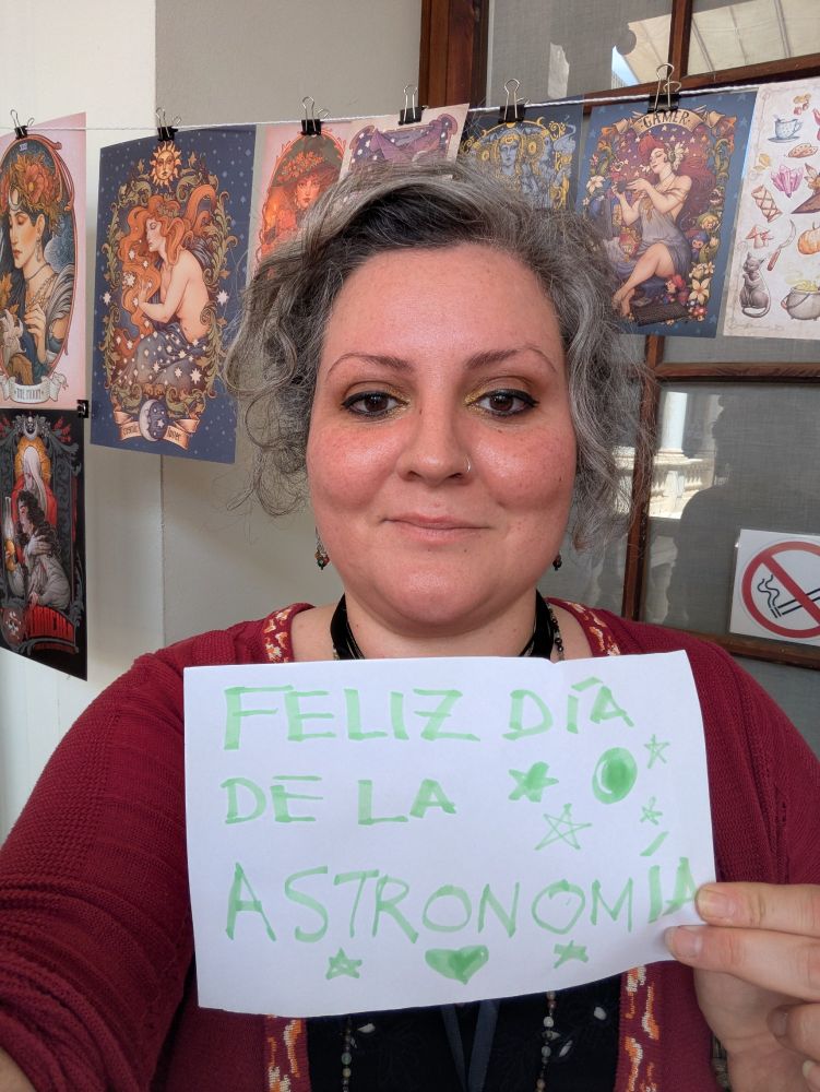 Señora cuarentona con un cartel pintado que pone FELIZ DÍA DE LA ASTRONOMÍA. Hay láminas colgadas detrás.