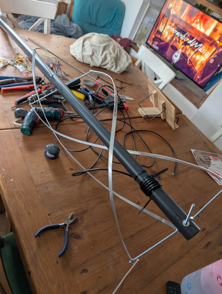  Mesa de madera llena de trastos y herramientas. Hay una antena en proceso montada en un tubo de pvc con cable coaxial y varillas roscadas. Ahora lleva unas pletinas de aluminio puestas. Al fondo está la tele encendida con una lista musical.