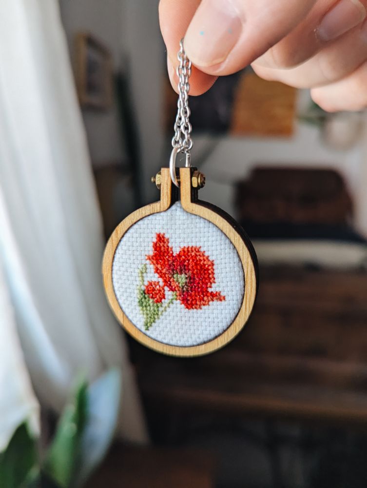 Un colgante de un bastidor de madera con un diseño de amapolas en punto de cruz. Al fondo, muebles y una ventana.