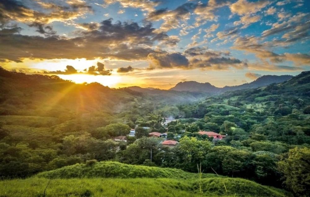 Image of village surrounded forested mountains. Sunset in the background.
