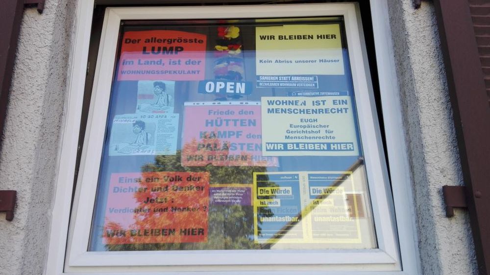 Ein Fenster mit Protestplakaten. "Wir bleiben hier" "der allergrößte Lump im Land, ist der Immobilienspekulant", "Friede den Hütten - Kampf den Palästen. Wir bleiben hier" ... Dazu Zeitungsartikel oder Flyer...