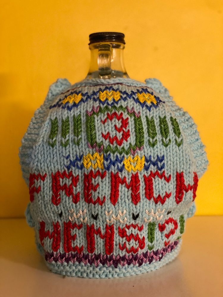 Front of a rooster-sized tabard displayed on a large clear glass jug, gaudily and colourfully decorated, reads: 3 FRENCH HENS!?!