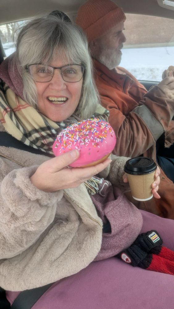 White-haired, bespectacled woman dressed for cold weather in the front passenger side of a car, holding a takeout coffee in one hand and a giant pink-frosted donut with sprinkles in the other. She’s grinning at the camera.