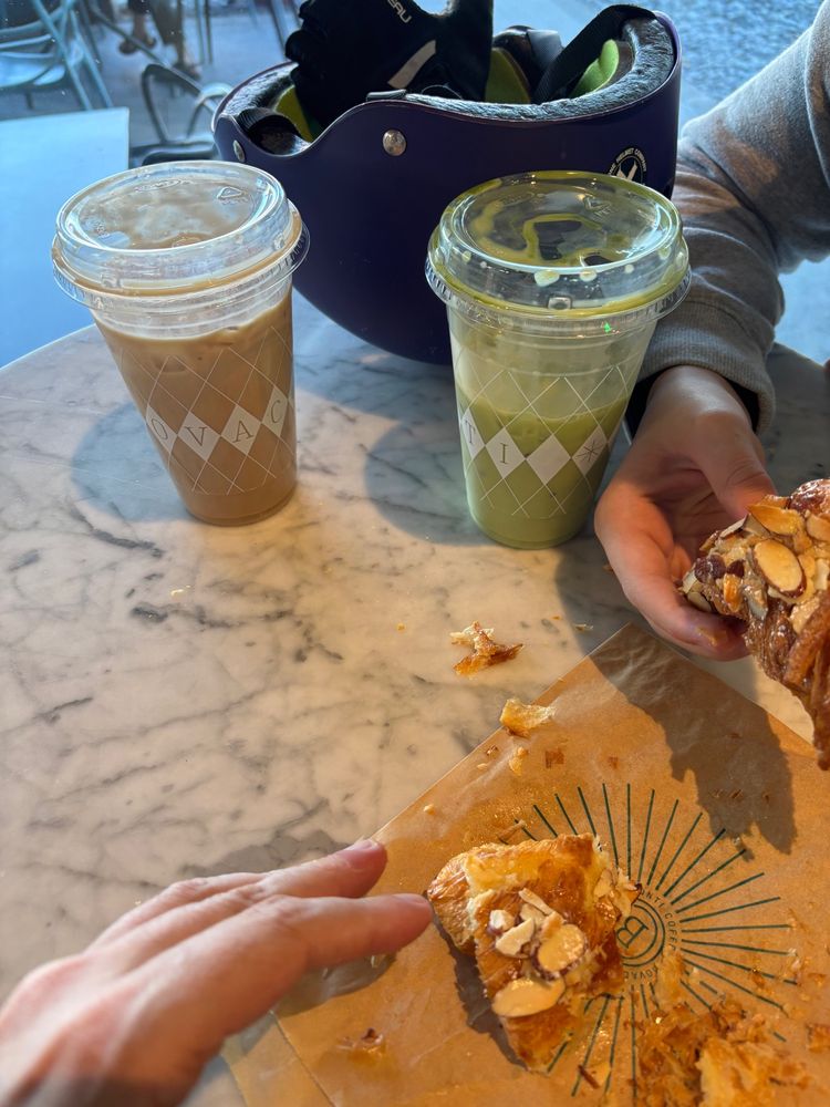Two iced coffee drinks a bike helmet and an almond croissant, being torn up by hands