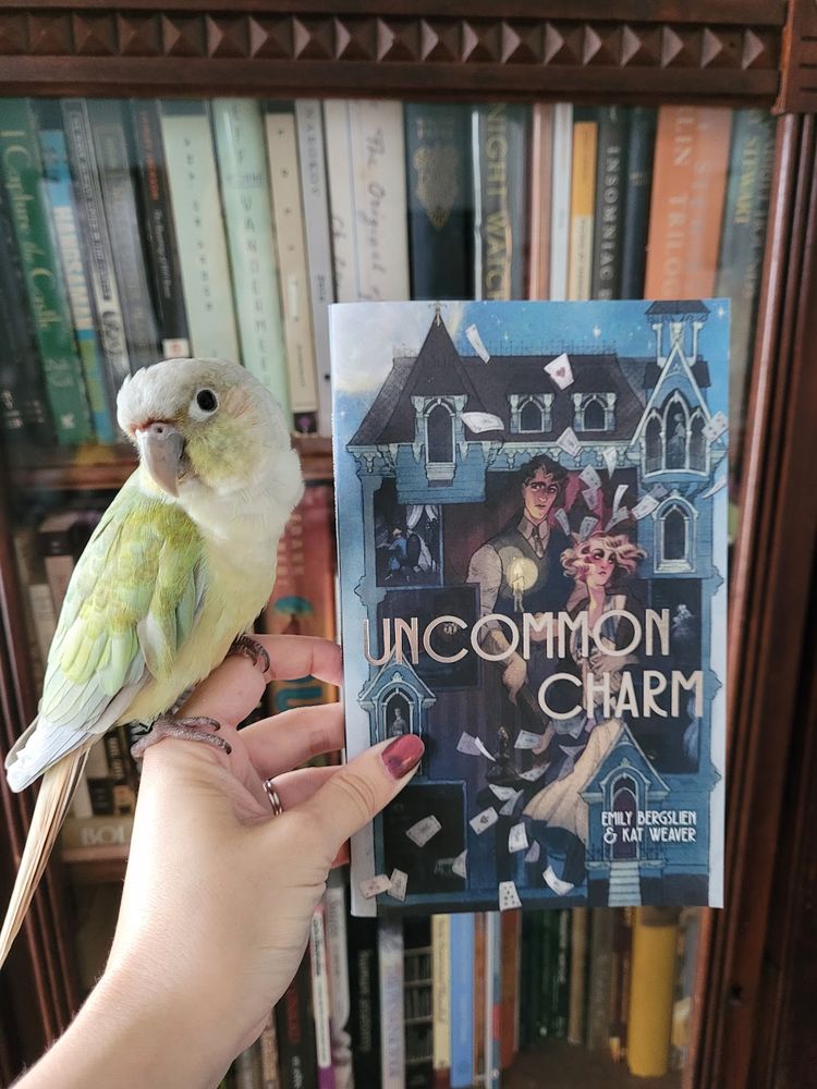 a handsome green cheeked conure poses beside a copy of Kat Weaver & Emily Berglisen's novella, Uncommon Charm