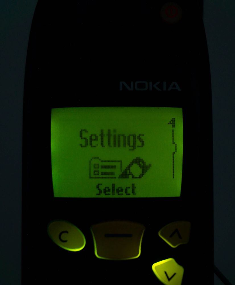 A photo of a Nokia 5190 powered on in the dark, highlighting its green-tinted frontlight.
The phone is displaying the "Settings" icon on the set of choices, with a 1-bit icon of a checklist folder and a Nokia 5110.

