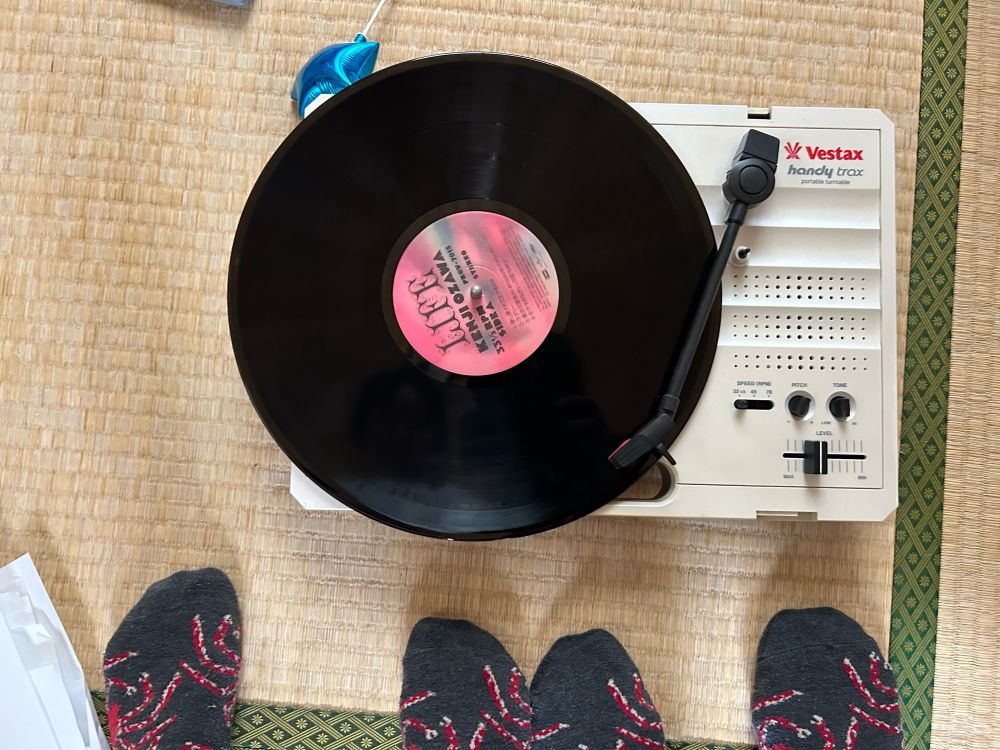 小沢健二「LIFE」アナログ盤がポータブルレコードプレイヤーの上に乗っている。
手前に靴下を履いた爪先が4つ（靴下の模様は黒地に赤いタカアシガニ）