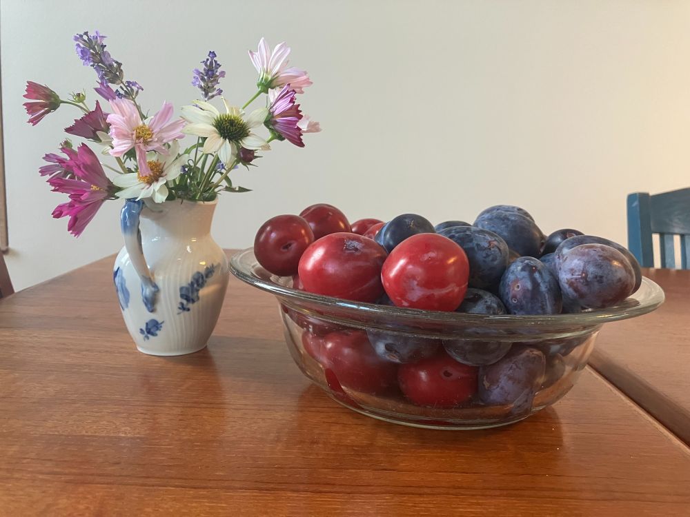 A blue and white vase on a wooden table holding white, pink, and purple flowers. Next to the vase on the table is a glass bowl holding plums.