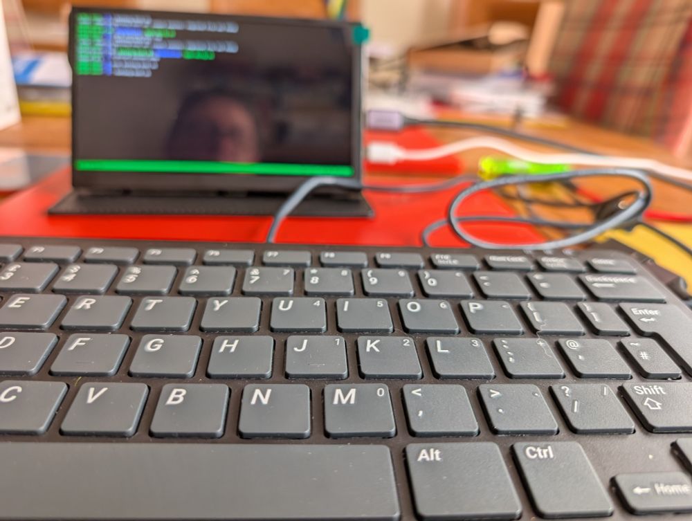 In the foreground, a computer keyboard. Behind it there's a small computer screen. The small green out-of-focus shape to the right is a Raspberry Pi Zero in a funky case