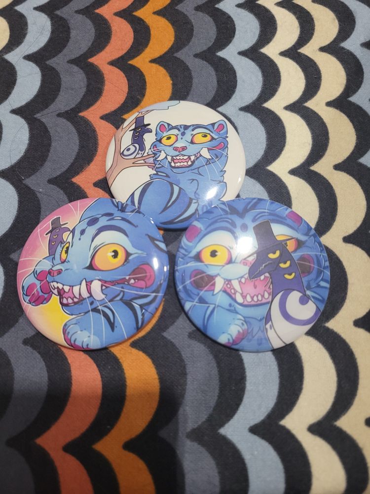 Three button pins are sitting on a patterned table cloth. They each feature different designs with the tiger and magpie from Kpop Demon Hunters